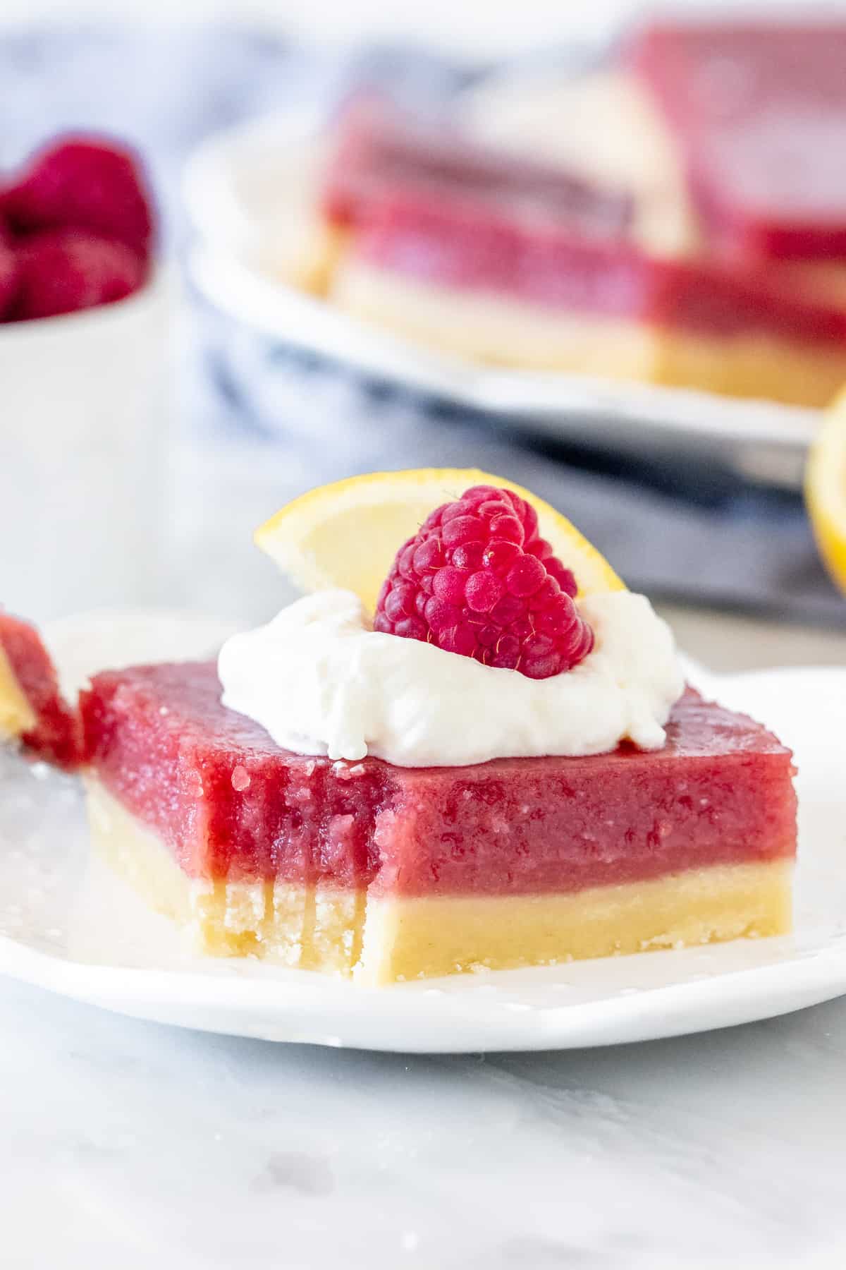 Lemon raspberry bar on a plate, topped with whipped cream and a berry. 