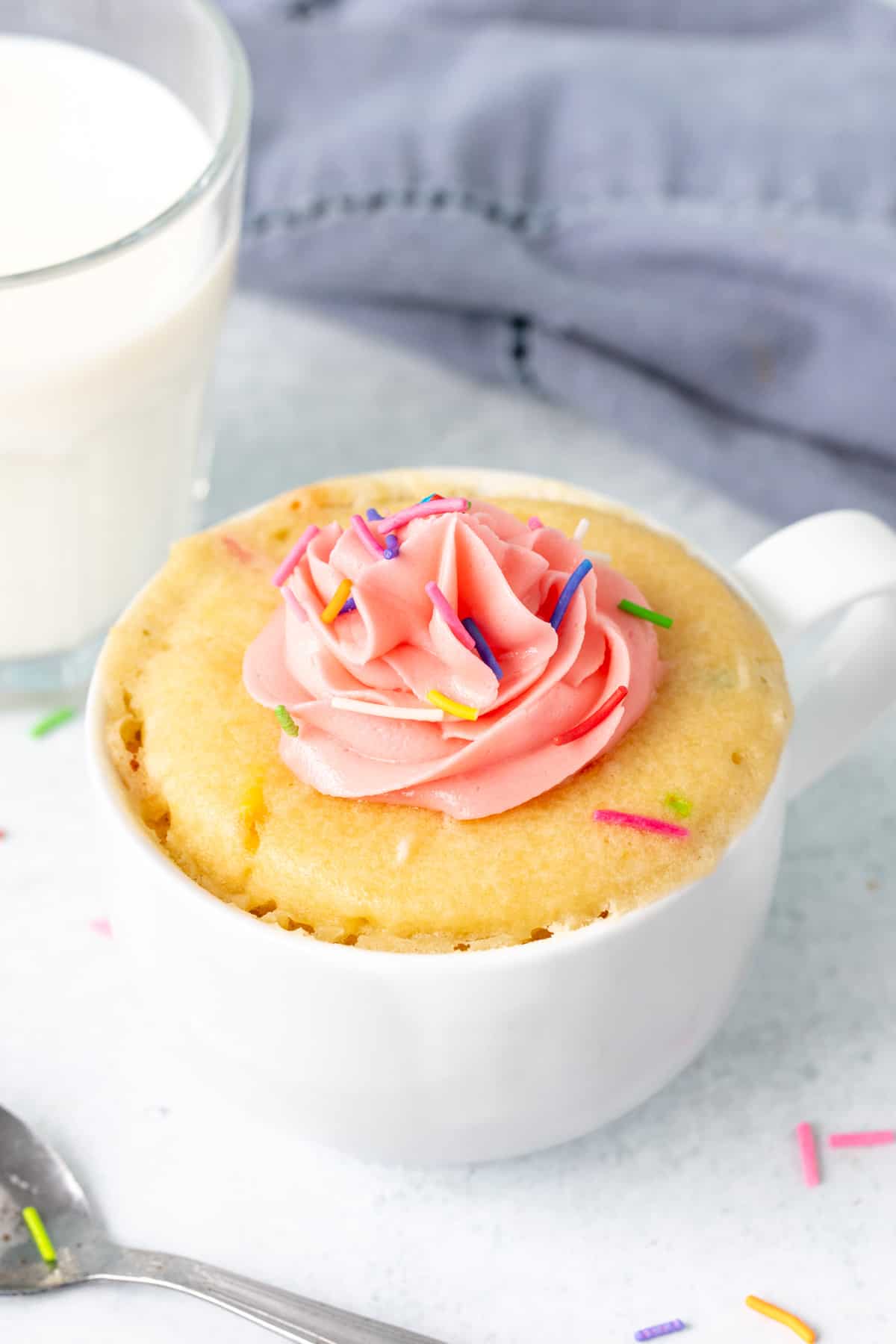 Vanilla mug cake topped with pink frosting and sprinkles with a glass of milk