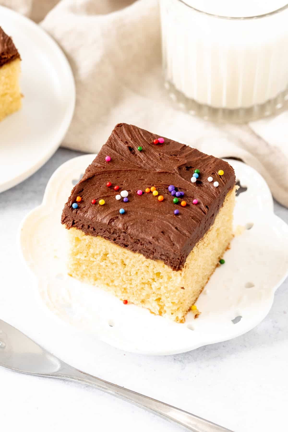 Square slice of golden yellow cake with chocolate frosting with a glass of milk