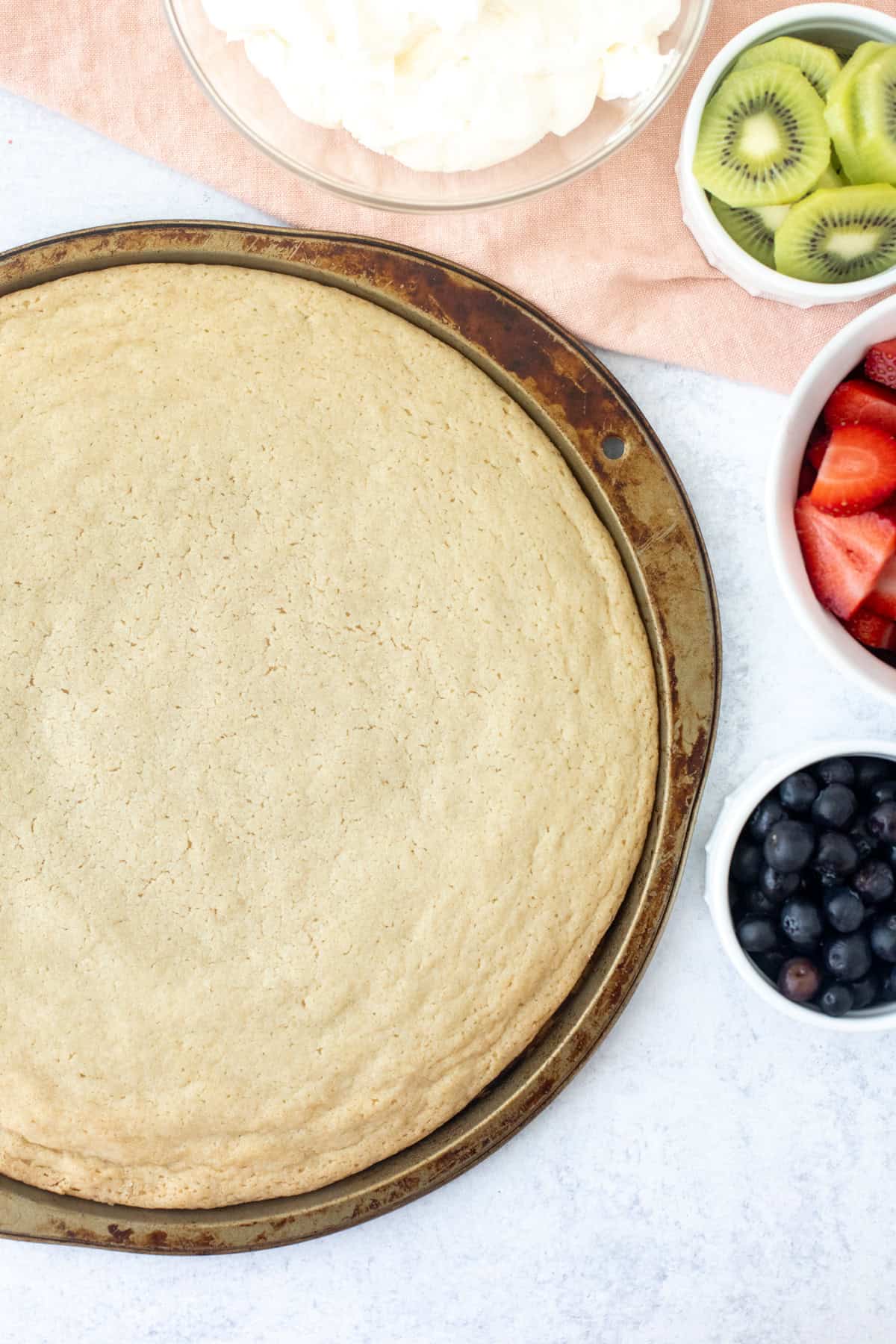 Sugar cookie made in a large pizza pan with bowl of whipped cream cheese frosting and bowls of fruit