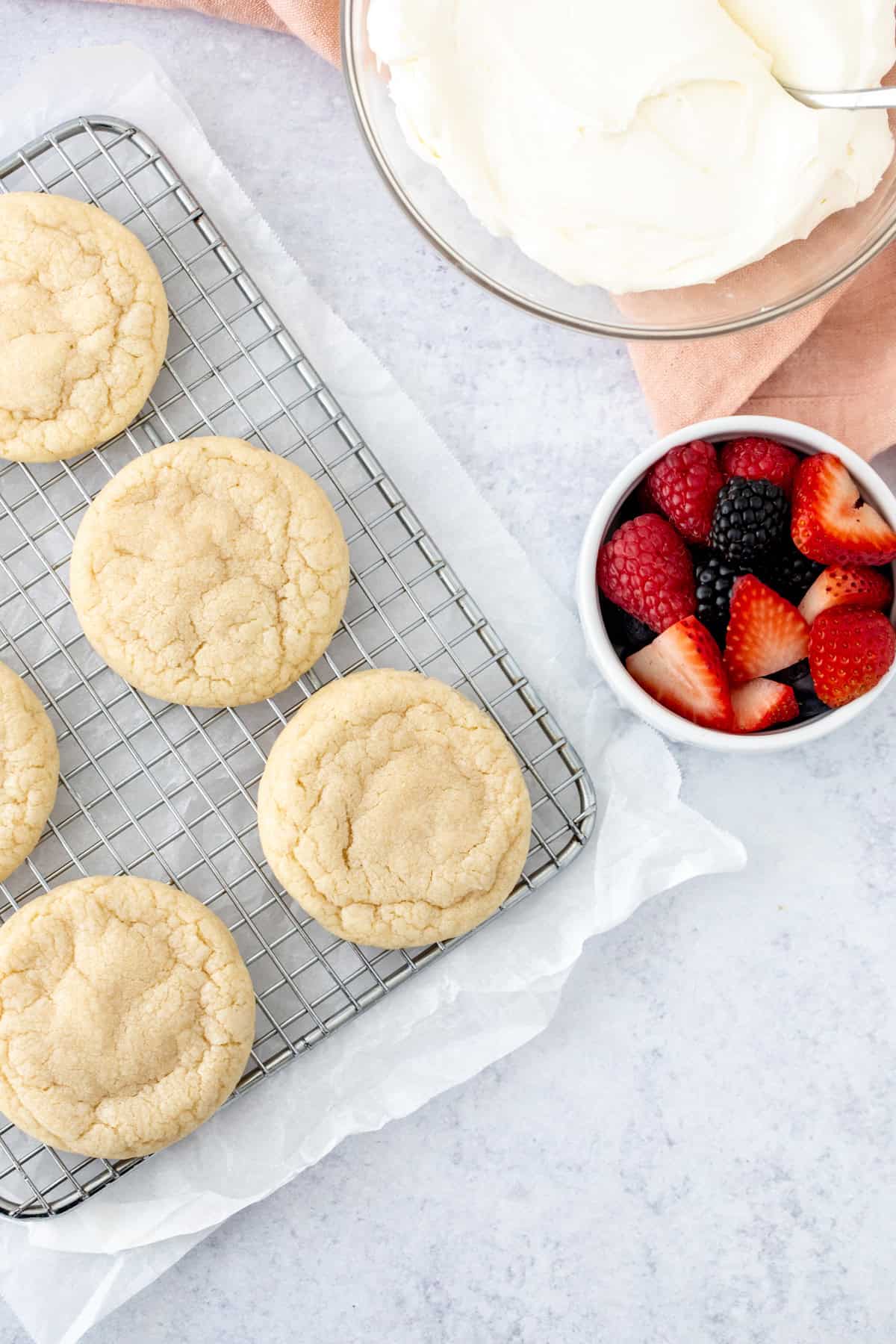 sugar cookies on wire rack with a bowl of whipped cream cheese frosting and berries