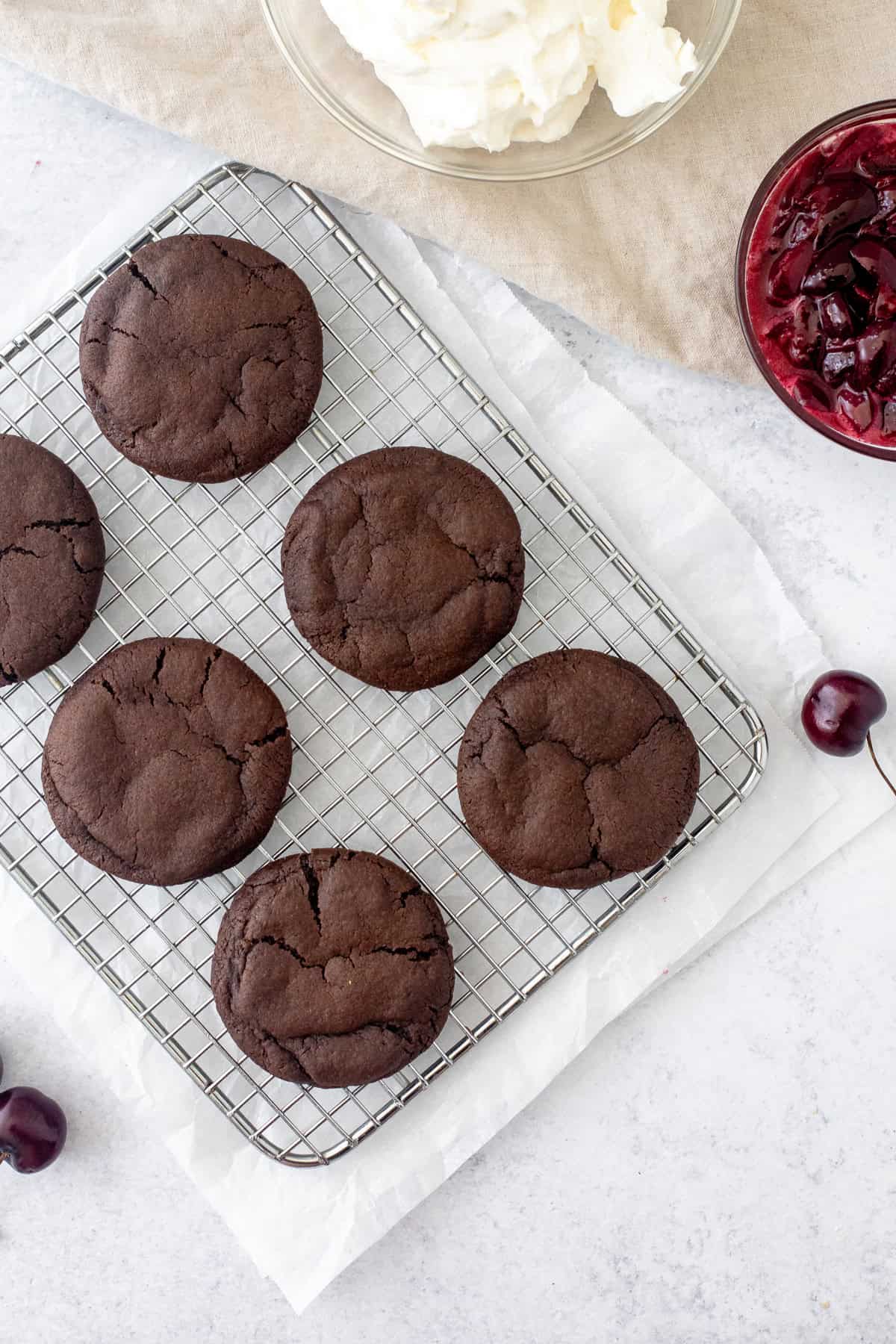 chocolate cookies on a cooling rack with bowl of whipped cream and bowl of cherry sauce